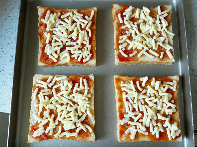 Slices of toast are coated with tomato sauce and sprinkled with mozzarella cheese.